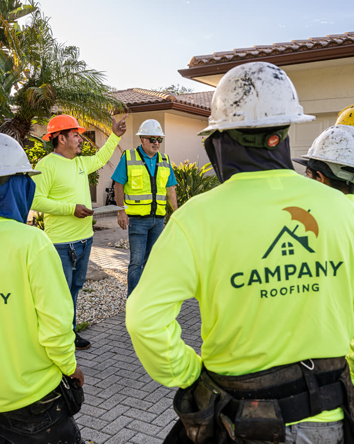 Campany Roofing crew in branded yellow shirts and hard hats gathered for a job briefing in front of a tile-roof home