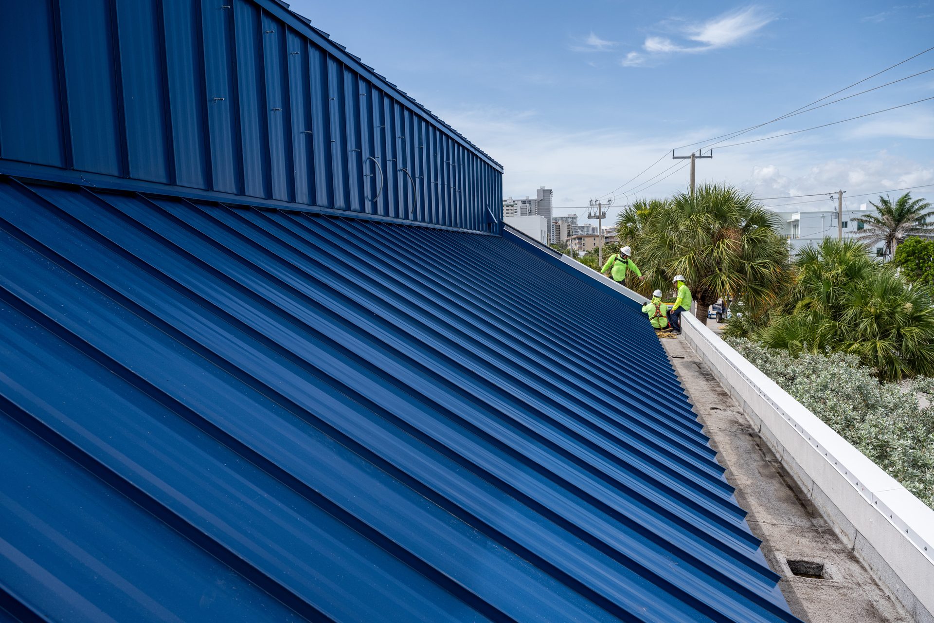 Campany Roofing crew installing a blue standing-seam metal roof overlooking a coastal South Florida town